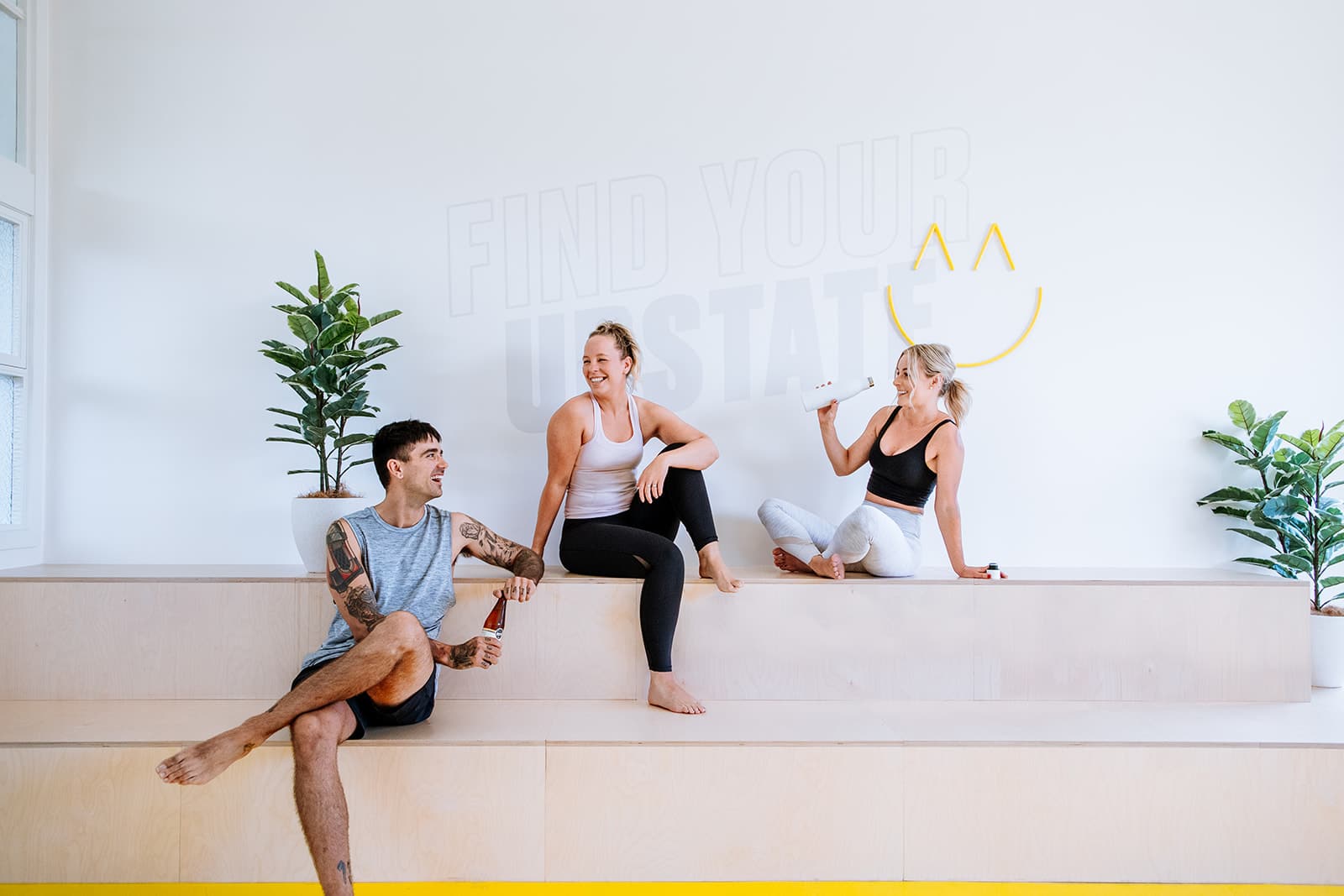 Three friends hang out happily at Upstate Studios in the communal area. Sleek warm wood and bright happy yellow hues create a motivating fitness studio environment.