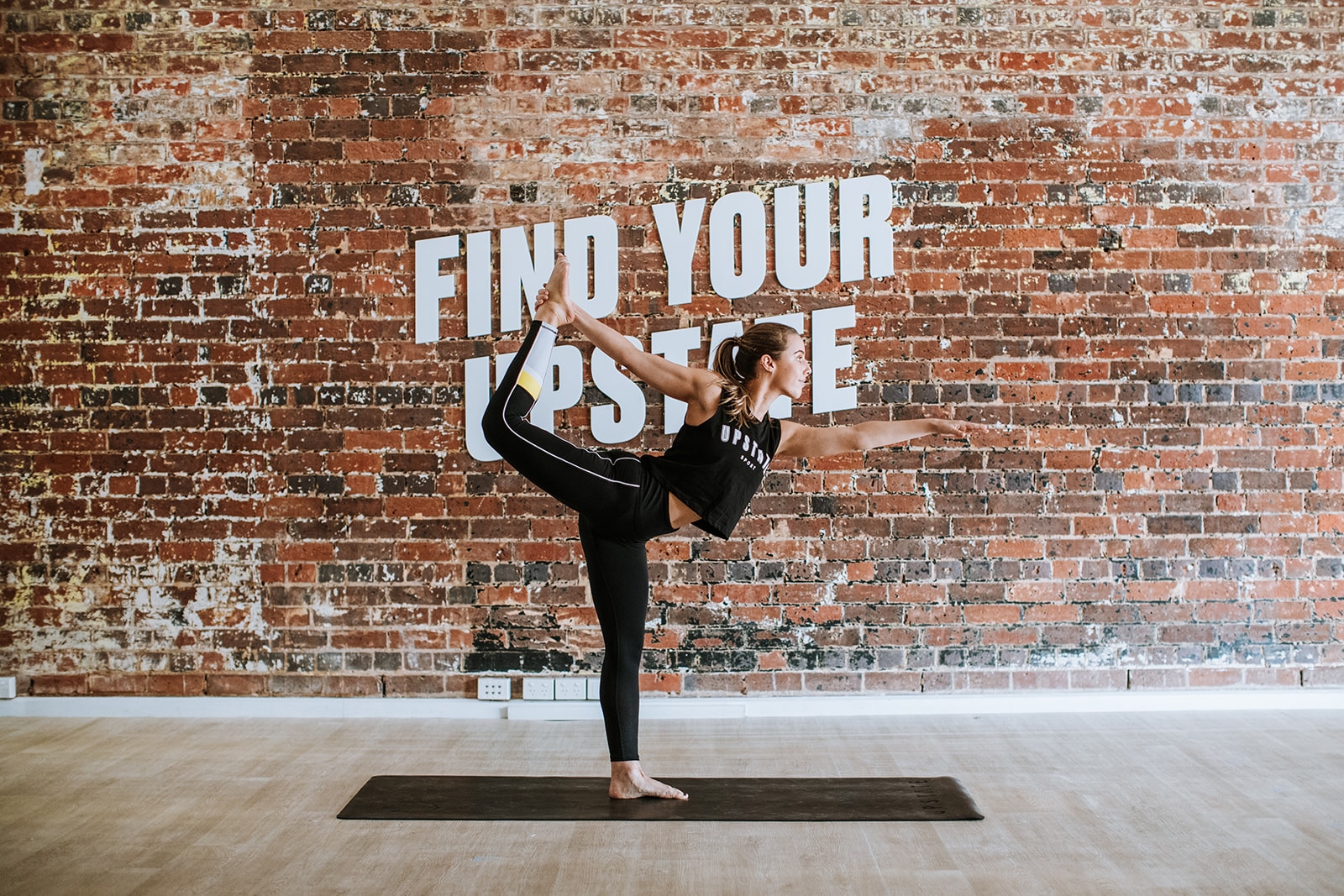 A woman in yoga pose, dancers pose, looks strong and graceful. She is doing yoga in the heated mat room of Upstate Studios. Behind her is a large brick wall with the words Find Your Upstate painted on in white.