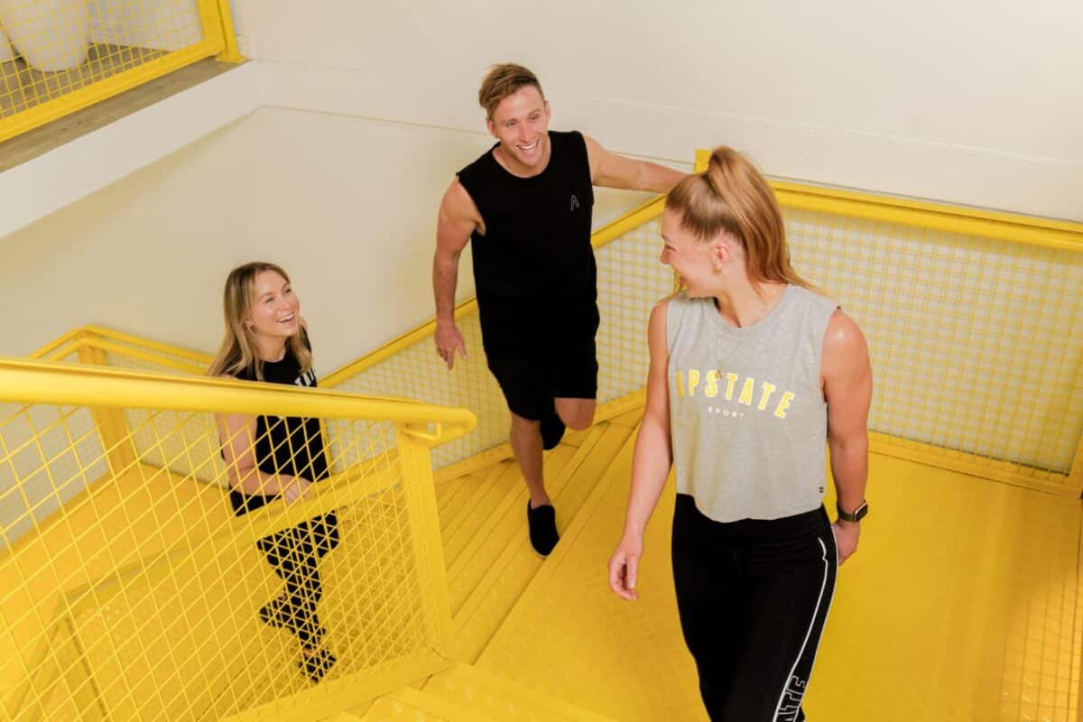 Upstaters smile at each other as they walk up happy yellow hued stairs to boxing studio.